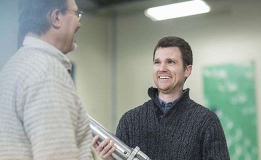 Person is discussing with a colleague while holding a machine part.